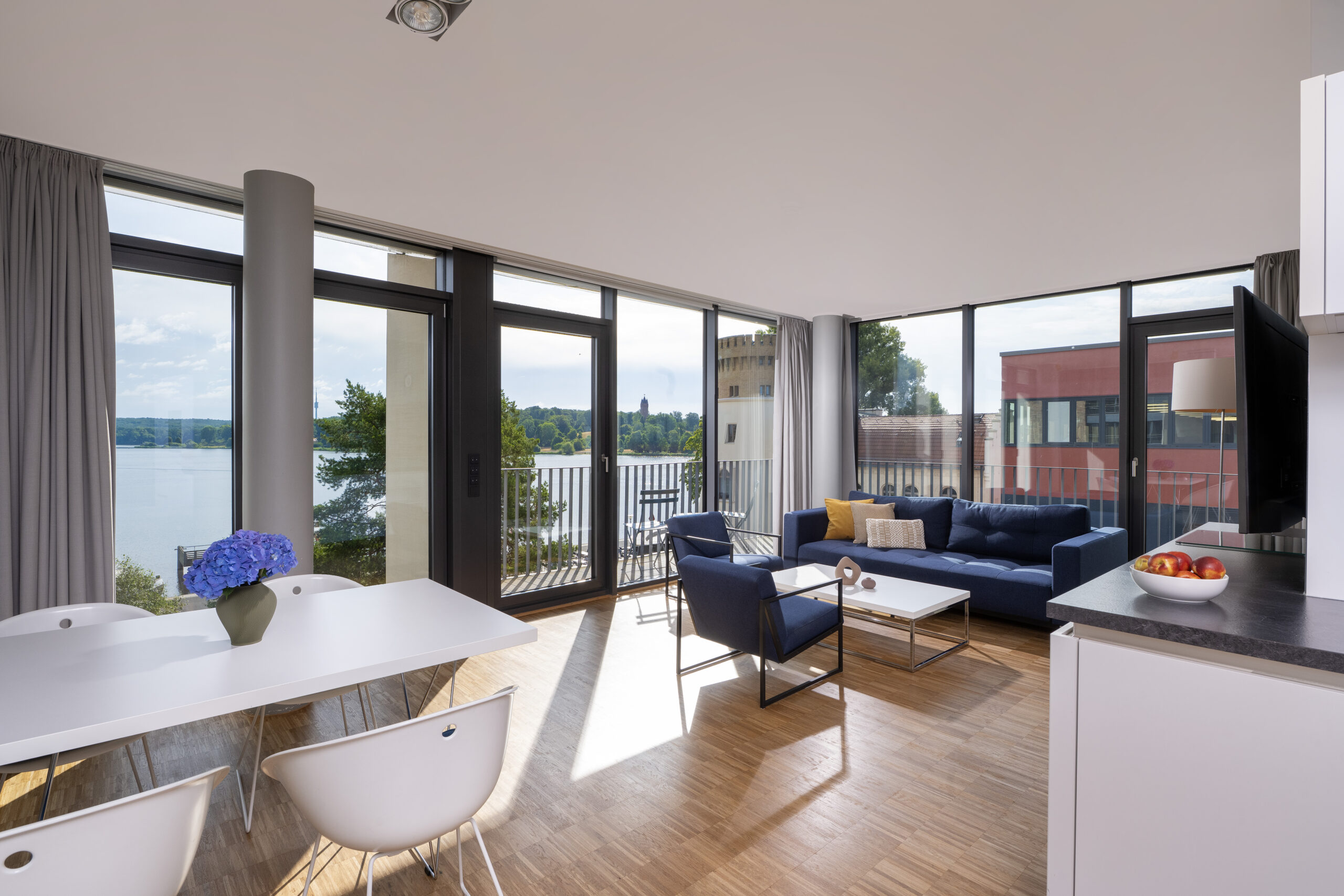 Ein geräumiges und modernes Corner-Apartment im Waveboard Hotel mit Panoramafenstern, die einen atemberaubenden Blick auf den See und das umliegende Stadtbild bieten. Das Apartment ist mit einem eleganten Sofa und einem gemütlichen Sessel ausgestattet, wodurch eine entspannte Atmosphäre entsteht.