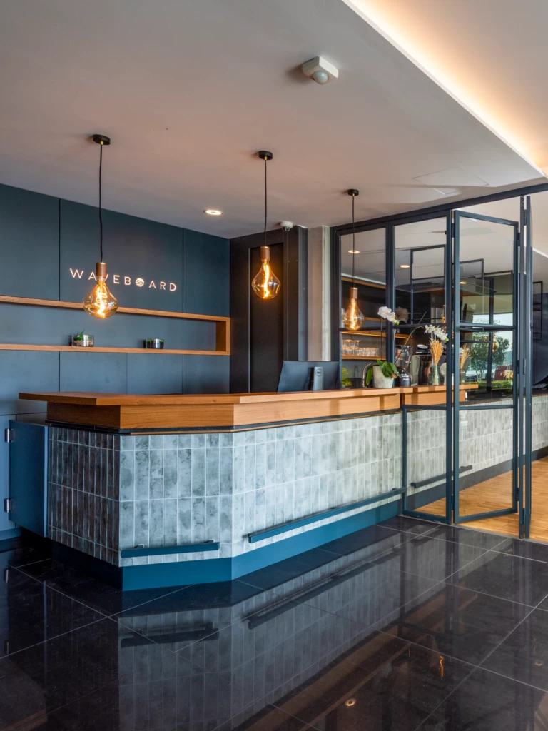 Moderne Rezeption im Waveboard Hotel Potsdam mit stilvoller Holz- und Fliesenverkleidung, hängenden Designerlampen und einem einladenden Empfangsbereich.