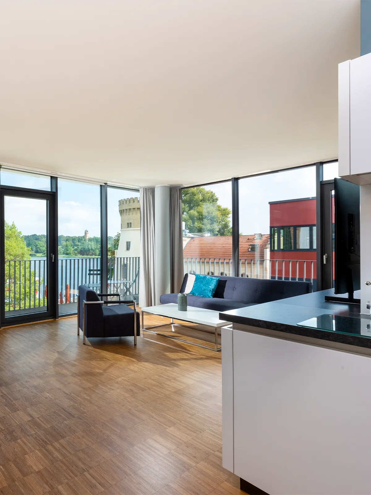 Heller Wohnbereich im Waveboard Hotel Potsdam mit bodentiefen Fenstern, stilvoller Einrichtung und Balkon mit Seeblick. Die offene Raumgestaltung verbindet Wohn- und Küchenbereich für ein modernes und komfortables Ambiente.
