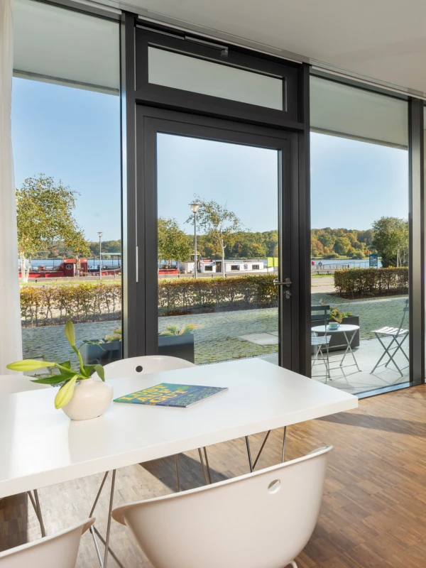 Erdgeschoss-Apartment im Waveboard Hotel mit einem modernen Tisch und Stühlen. Große Fenster bieten einen direkten Blick auf den Außenbereich und den Templiner See, ideal für entspannte Momente.